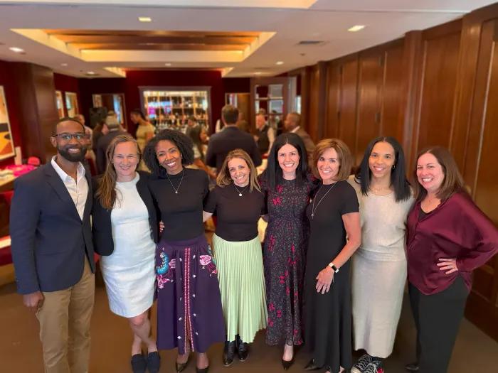 Smiling group shot of eight Bright Pink board members standing in a row together. They are wearing business casual and appear to be in the lobby of a restaurant.