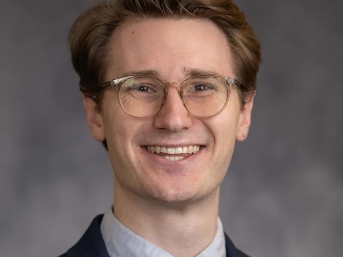 White male with dark red hair and glasses, wearing a dark suit jacket, blue shirt, and dark tie.