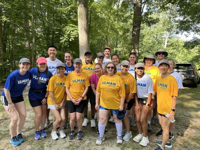 A group of 19 Cancer to 5K participants wearing a mix of maize, blue and white shirts pose outdoors in front of trees