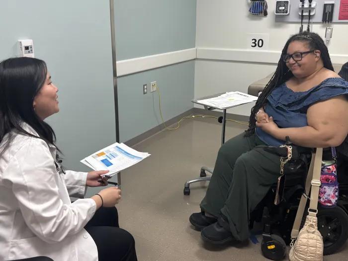 A medical student with a standardized patient who uses a wheelchair have a conversation in an exam room