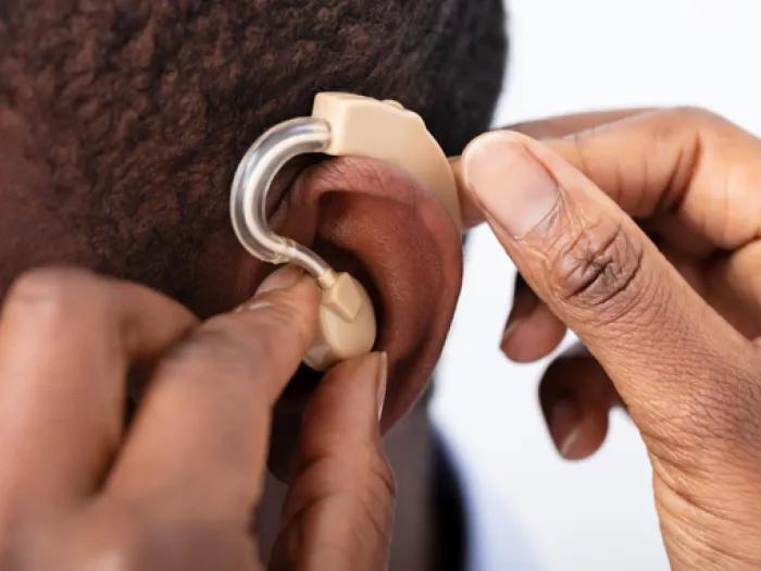 A close-up of a person’s ear as they insert a beige behind-the-ear hearing aid, using both hands to position the device properly.