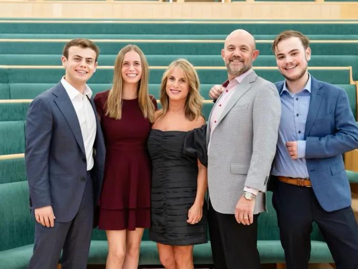 Smiling family photo of 5 members of the Kripke family. They appear to be dressed for a semi-formal event and are standing in an open space with green-cushioned seating visible behind them.