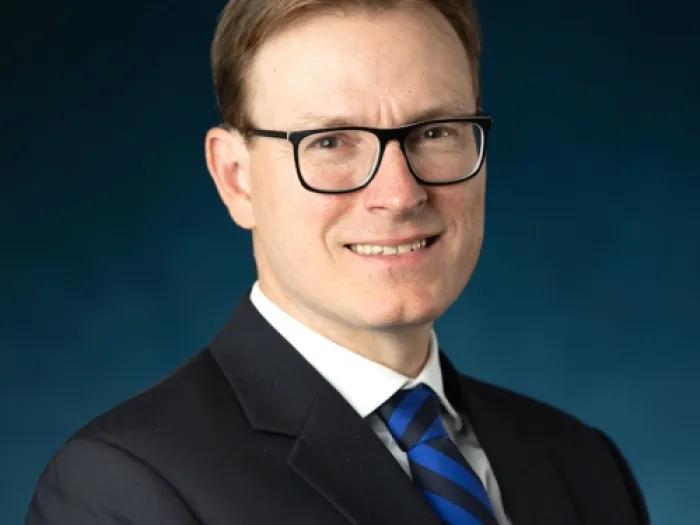 White male with dark glasses, wearing a black suit jacket and a blue and black tie