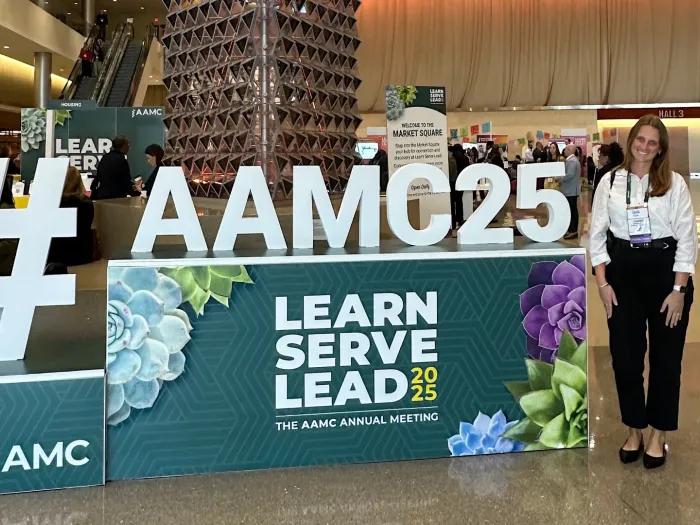 A woman stands next to a display in a conference center that reads #AAMC25 Learn Serve Lead
