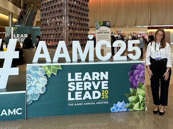 A woman stands next to a display in a conference center that reads #AAMC25 Learn Serve Lead