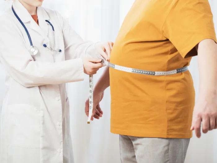 a doctor with a lab coat mesuring an overweight person in a yellow shirt using a tape measure around their midsection