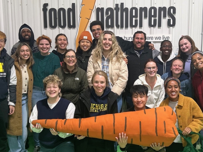 A group of people pose in front of a corrugated metal wall with a "food gatherers" sign and a large carrot graphic. Several individuals are wearing University of Michigan Medical School apparel, and four people in front hold a large plush carrot.