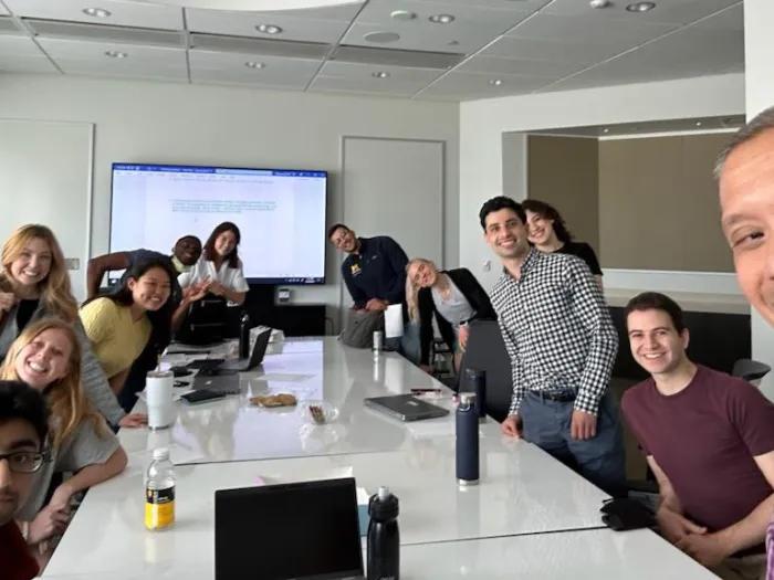 Group of people gathered around a large conference table in a modern classroom, with laptops, water bottles, and snacks on the table. A large screen displays a document on the wall, and the group appears to be smiling and interacting casually.