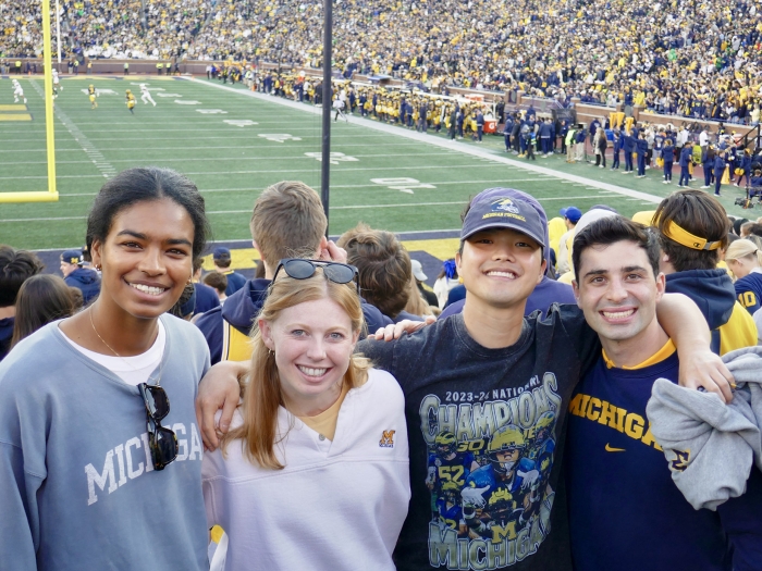 A group of friends wearing University of Michigan apparel pose together in the stands at Michigan Stadium during a football game, with the field, goalposts, and a large crowd of fans visible in the background.