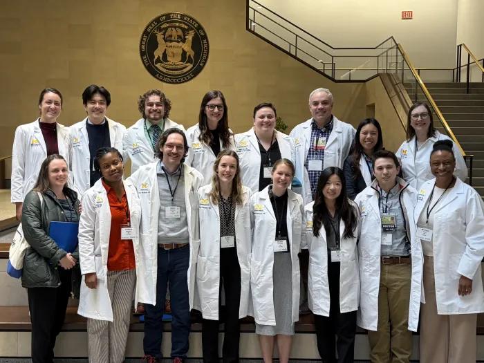 Sixteen people stand in two rows looking at the camera and smiling. Most are wearing white physician coats. The State of Michigan seal is on the wall behind them. 
