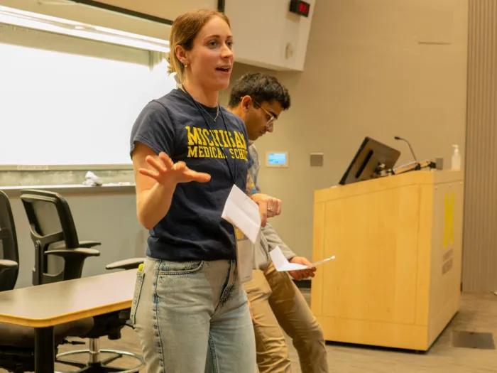 Two people stand at the front of a lecture hall, one wearing a Michigan Medical School t-shirt and gesturing, near a podium with the University of Michigan logo.