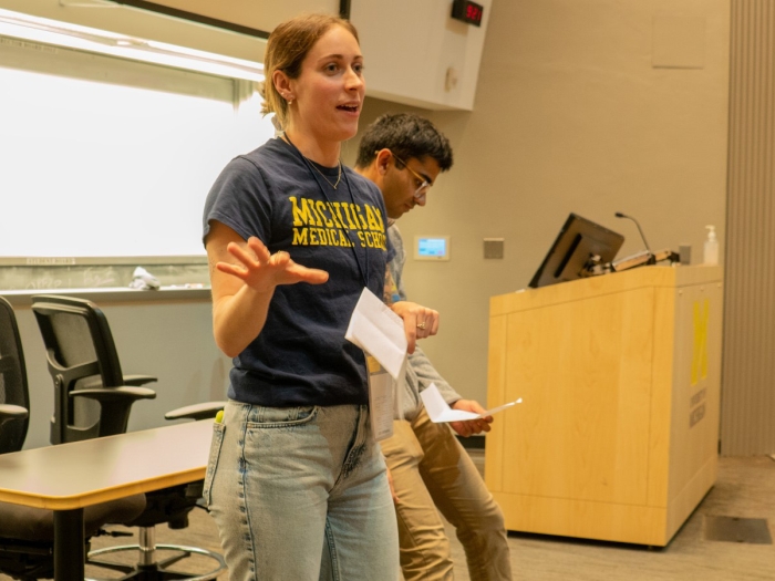 Two people stand at the front of a lecture hall, one wearing a Michigan Medical School t-shirt and gesturing, near a podium with the University of Michigan logo.