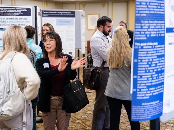 Attendees network at a poster session in a conference hallway, standing in small groups and discussing research displayed on large poster boards and pull-up banners.