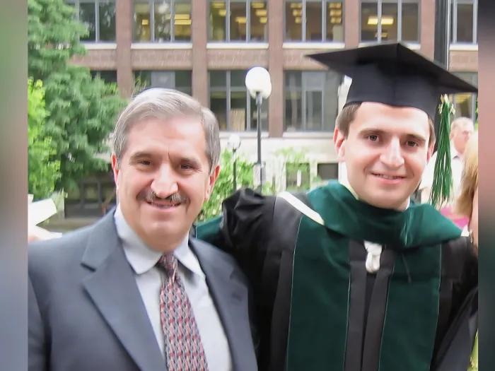Ahmad and Habib Rahbar on Habib's Medical School graduation day. They are smiling and standing together in front of a U-M building, and Habib is wearing a black graduation gown and mortarboard with a green tassel and sash.