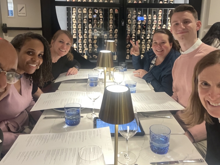 A group of people sitting around a rectangular dining table set with blue water glasses, wine glasses, and menus. Three gold-colored lamps are placed along the center of the table. In the background, there is a glass wall displaying numerous wine bottles. The atmosphere appears to be that of a modern, upscale restaurant.