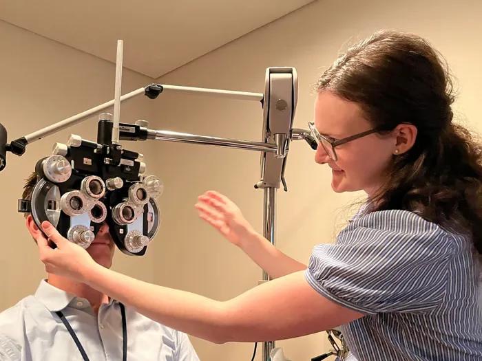 Resident adjusts a phoropter during an eye exam while another resident looks through the device, demonstrating clinical training in vision testing.