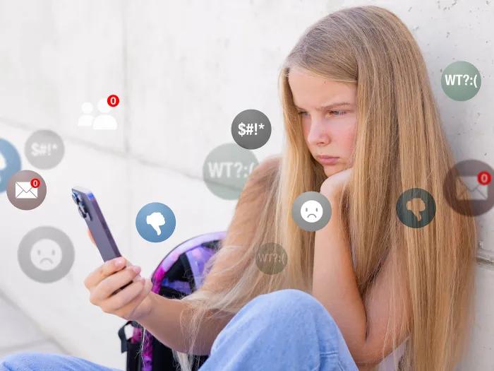Teen girl sitting on floor next to her bed looking at a smartphone, with icons from social media apps layered over her as she frowns