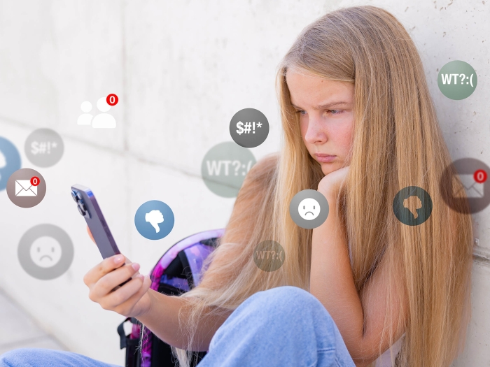 Teen girl sitting on floor next to her bed looking at a smartphone, with icons from social media apps layered over her as she frowns