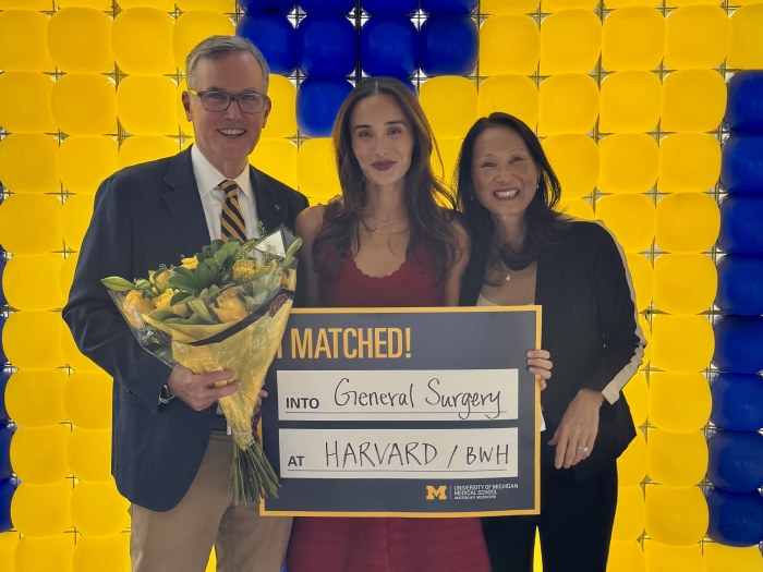 Alt text: **Three people pose in front of a maize-and-blue balloon wall; the person on the left holds a bouquet, and they hold a sign reading “MATCHED! into General Surgery at HARVARD / BWH” with University of Michigan Medical School branding.