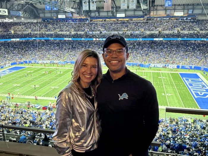 Two people pose in the foreground at an indoor Detroit Lions football game, with a packed stadium and the field visible behind them; one wears a shiny silver jacket and the other a black Lions pullover and cap.