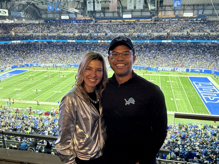Two people pose in the foreground at an indoor Detroit Lions football game, with a packed stadium and the field visible behind them; one wears a shiny silver jacket and the other a black Lions pullover and cap.