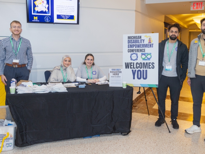 Five conference staff wearing name badges stand and sit around a registration table in a building lobby. A large sign on an easel reads “Michigan Disability Empowerment Conference Welcomes You,” with a smaller sign that says “Register Here.”