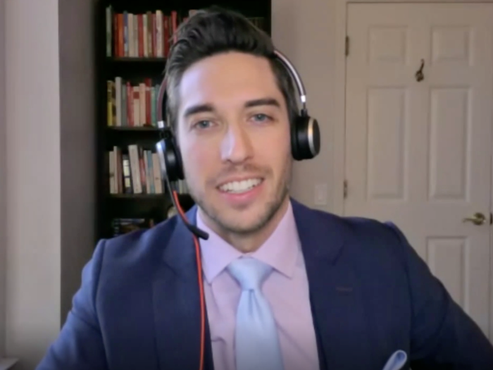 Person in a navy suit with a light pink shirt and light blue tie wears a headset while seated at a desk in a home office; a bookshelf and closed double doors are in the background, with a window on the left.