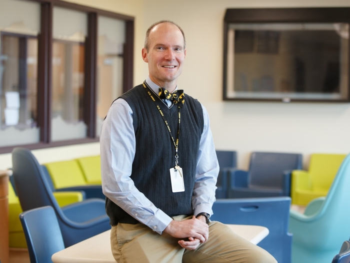 Chris Thurstone smiling and sitting on a desk in a brightly colored room