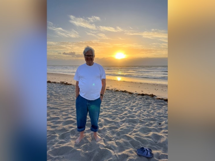 An older man stands on a white sand beach with the sun setting into the ocean behind him. He is wearing a white t-shirt and jeans. 