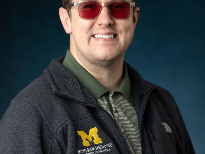 White man with dark brown hair, amber eyeglass frames, and a dark grey U-M logo sweater