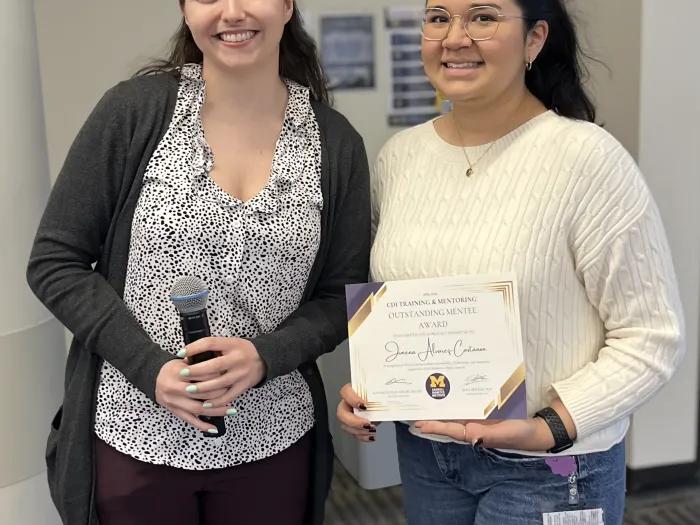 Two women stand side by side at the CDI Training and Mentoring Awards event, smiling at the camera. The woman on the left holds a microphone, while the woman on the right holds an Outstanding Mentee Award certificate. The photo was taken indoors against a neutral background with a dark blue border surrounding the image.
