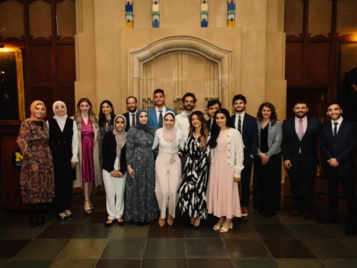 A group of about 18 people pose for a formal photo in a historic hall with a large wooden door and portraits on the walls, wearing a mix of suits and dresses, some with hijabs.