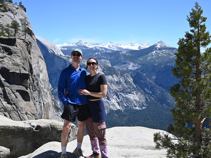 Two hikers pose together on a rocky overlook with a pine tree nearby and snow-capped mountains in the distance.