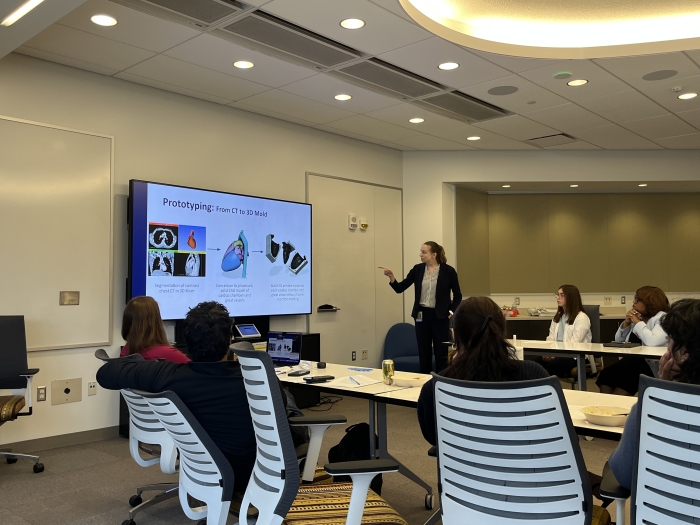 A woman in a dark suit presenting to a group sitting at a u-shaped table. 