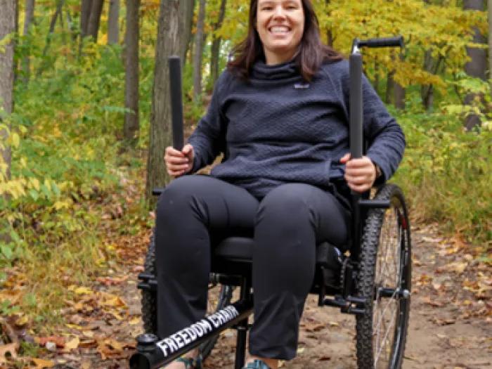 A person using an all-terrain wheelchair sits on a wooded trail surrounded by yellow fall foliage.