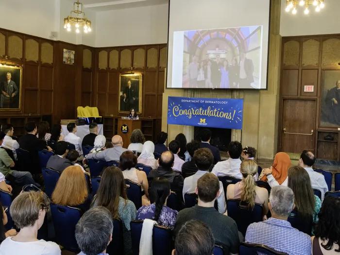 Woman standing at podium speaking to a large seated group of people attending a graduation event