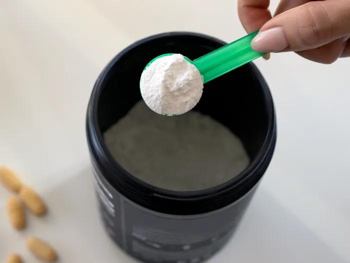 A hand holding a scoop of protein powder hovering over a cup of water