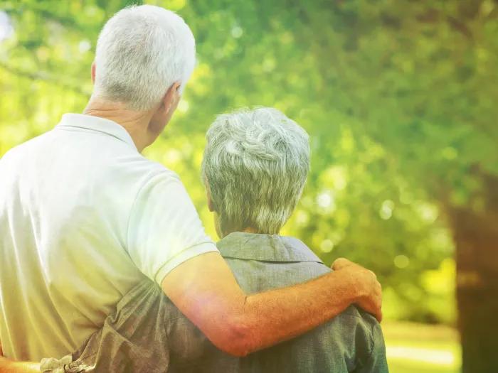 A man with his arm around a women's shoulder, both seniors and shown from the back, staring off into a forest.