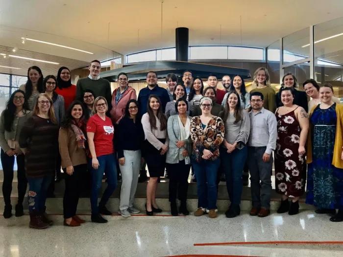 A group of about 25 people pose for a photo inside a modern building lobby with large windows and overhead lighting. 