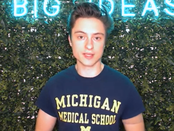 A person wears a navy “Michigan Medical School” T-shirt while standing in front of a leafy green wall with a blue neon sign partially reading “BIG IDEAS.”