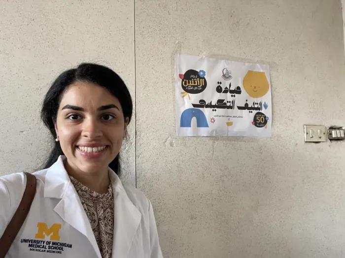 A person wearing a University of Michigan Medical School white coat smiles for a selfie beside a health education poster with Arabic text on a wall.