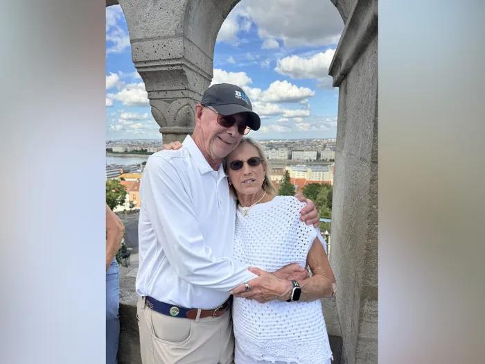 Bill and Sally Martin stand affectionately with their arms around each other. They appear to be in a high open tower above a city.