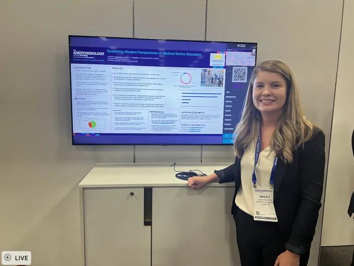 A person wearing a conference badge stands smiling beside a digital research poster titled “Examining Student Perspectives on Medical Device Education” displayed on a large screen.