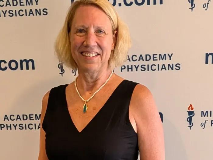 Women with chin-length blond hair, wearing a black, V-neck blouse and necklace.