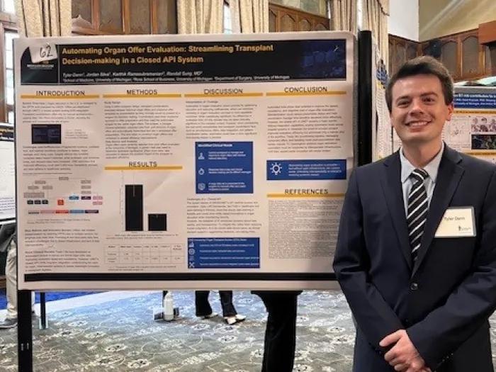 A person in a suit stands smiling beside a research poster titled “Automating Organ Offer Evaluation: Streamlining Transplant Decision-making in a Closed API System” at an indoor academic poster session.