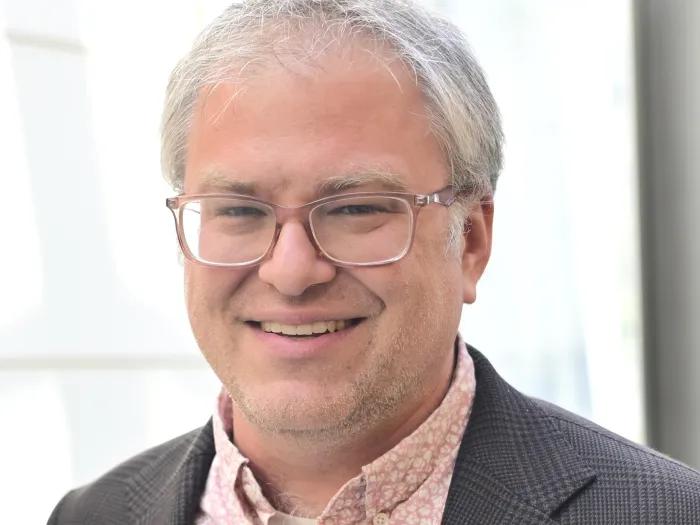 Close-up portrait of a smiling man with short gray hair and glasses, wearing a pink patterned shirt and dark blazer against a bright, softly blurred background.