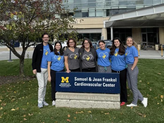 Seven research team members wearing matching blue and gray shirts pose outdoors behind a University of Michigan Health sign for the Samuel & Jean Frankel Cardiovascular Center.