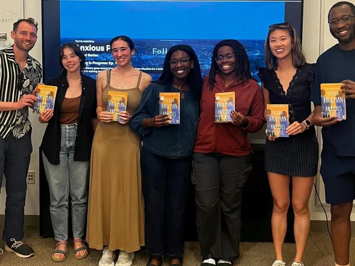 Seven smiling medical students stand together in a classroom, six holding copies of Fredrik Backman’s novel Anxious People, with a presentation screen behind them.