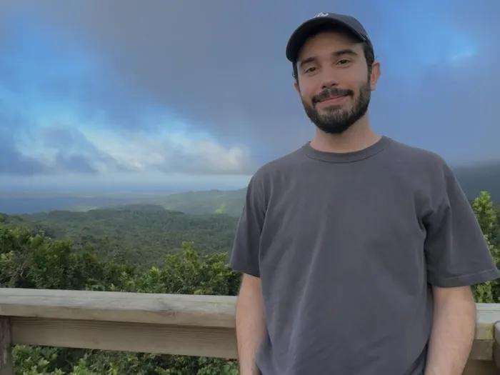 Med student Noah Carey, a smiling bearded man in a dark cap and T-shirt stands at a scenic overlook above lush green mountains and the ocean beneath a cloudy blue sky.