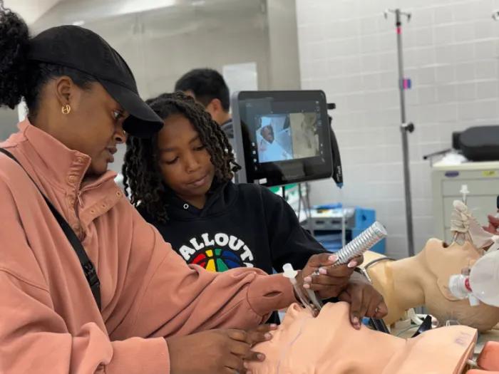 An adult guides a child inserting a breathing tube into an airway-training mannequin in a medical simulation room. 